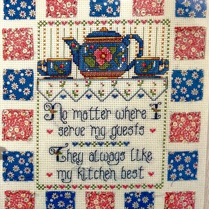 May include: Cross-stitch sampler featuring a blue teapot and two teacups on a shelf, with the text "No matter where I serve my guests, they always like my kitchen best." The sampler is framed by alternating blue floral and red floral squares.