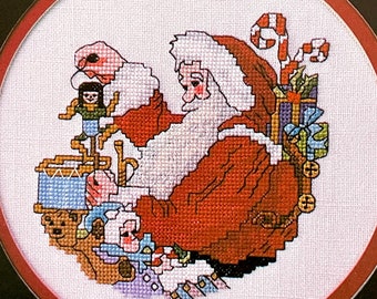 May include: A cross-stitch design featuring Santa Claus, dressed in a red suit with a white beard and hat, surrounded by toys and gifts. The design includes a toy soldier, teddy bear, and candy canes, all within a circular frame.