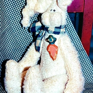 May include: A plush, cream-colored bunny with long ears and a blue and white checkered scarf. The bunny has a carrot-shaped applique and is sitting on a checkered surface. A small pile of red berries is at the bottom.