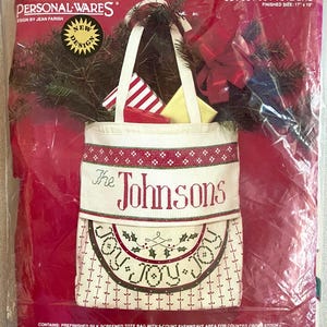 May include: A Christmas cross-stitch tote bag kit. The bag is white with a red and green border and the name "The Johnsons" stitched across the front. The bag is filled with gifts and greenery. Finished size is 17 x 19 inches.