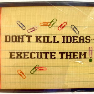 May include: Cross-stitched artwork on a notepad design. The text reads "DON'T KILL IDEAS... EXECUTE THEM" in dark brown letters. Colorful paper clips in red, green, orange, and purple are scattered across the notepad.
