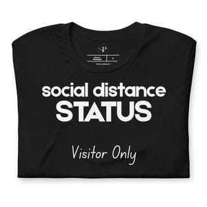 May include: Black t-shirt with the words "social distance STATUS" in large white block letters. Below, in a cursive font, it reads "Visitor Only". The shirt is folded.