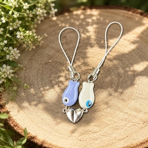 May include: Two phone charms featuring fish-shaped pendants in light blue and white, each with a dark blue and white evil eye detail. The pendants are connected to a central silver-toned metal heart charm and include thin silver-toned attachment loops.