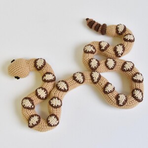 Könnte beinhalten: Ein gehäkeltes Schlangenspielzeug in Beige mit braunen und cremefarbenen Akzenten. Die Schlange hat einen abgerundeten Kopf mit kleinen schwarzen Augen und einen gebogenen Körper. Der Körper ist mit gleichmäßig verteilten, kreisförmigen braunen und cremefarbenen Mustern verziert. Das Spielzeug ist weich und sicher für Kinder.