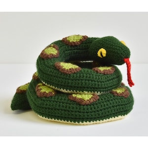 May include: Crocheted snake toy in shades of green, brown, and yellow. The snake is coiled, with a red tongue extending from its mouth. The toy is handmade and sits on a white surface.