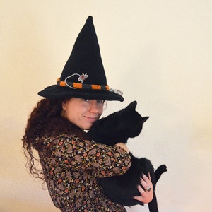 May include: A woman wearing a black witch hat with orange and grey trim holds a black cat in her arms.