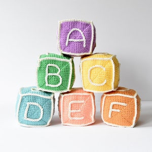 Op de afbeelding: Een set van zes gehaakte alfabetblokken, elk met een andere letter van A tot en met F. De blokken zijn in een piramidevorm gestapeld, met de bovenste blok paars, de volgende twee blokken groen en geel, en de onderste drie blokken blauw, roze en oranje.