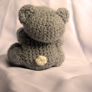 May include: A gray crocheted teddy bear sitting on a white fabric surface. The bear has a white button nose and is facing away from the camera.