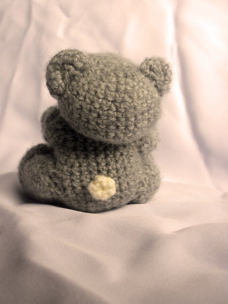 May include: A gray crocheted teddy bear sitting on a white fabric surface. The bear has a white button nose and is facing away from the camera.