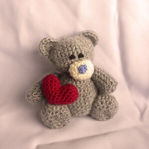 May include: A gray crocheted teddy bear holding a red heart. The bear has a white nose with a blue button.