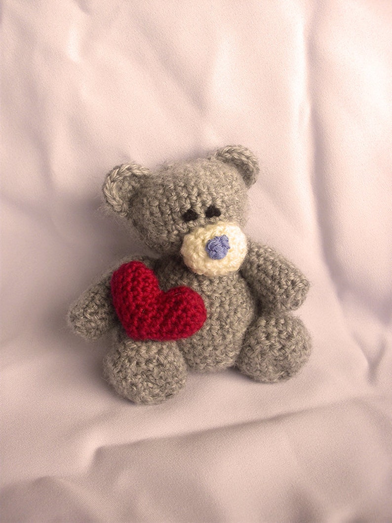 May include: A gray crocheted teddy bear holding a red heart. The bear has a white nose with a blue button.