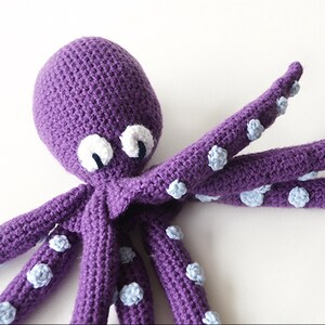 May include: A handmade purple crochet octopus with white and blue accents. The octopus has a rounded head, large white eyes with black pupils, and eight long tentacles. The tentacles are adorned with small, light blue suckers.