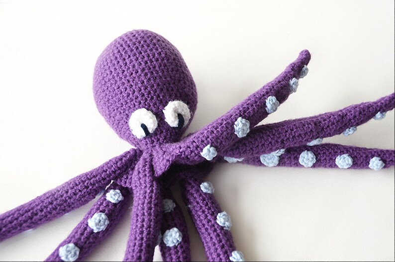May include: A handmade purple crochet octopus with white and blue accents. The octopus has a rounded head, large white eyes with black pupils, and eight long tentacles. The tentacles are adorned with small, light blue suckers.
