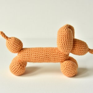 May include: A crocheted stuffed toy shaped like a dachshund, with a light orange color. The toy has a long body and a curled tail.