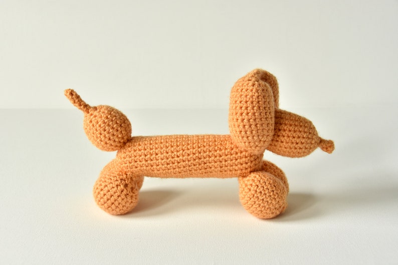 May include: A crocheted stuffed toy shaped like a dachshund, with a light orange color. The toy has a long body and a curled tail.