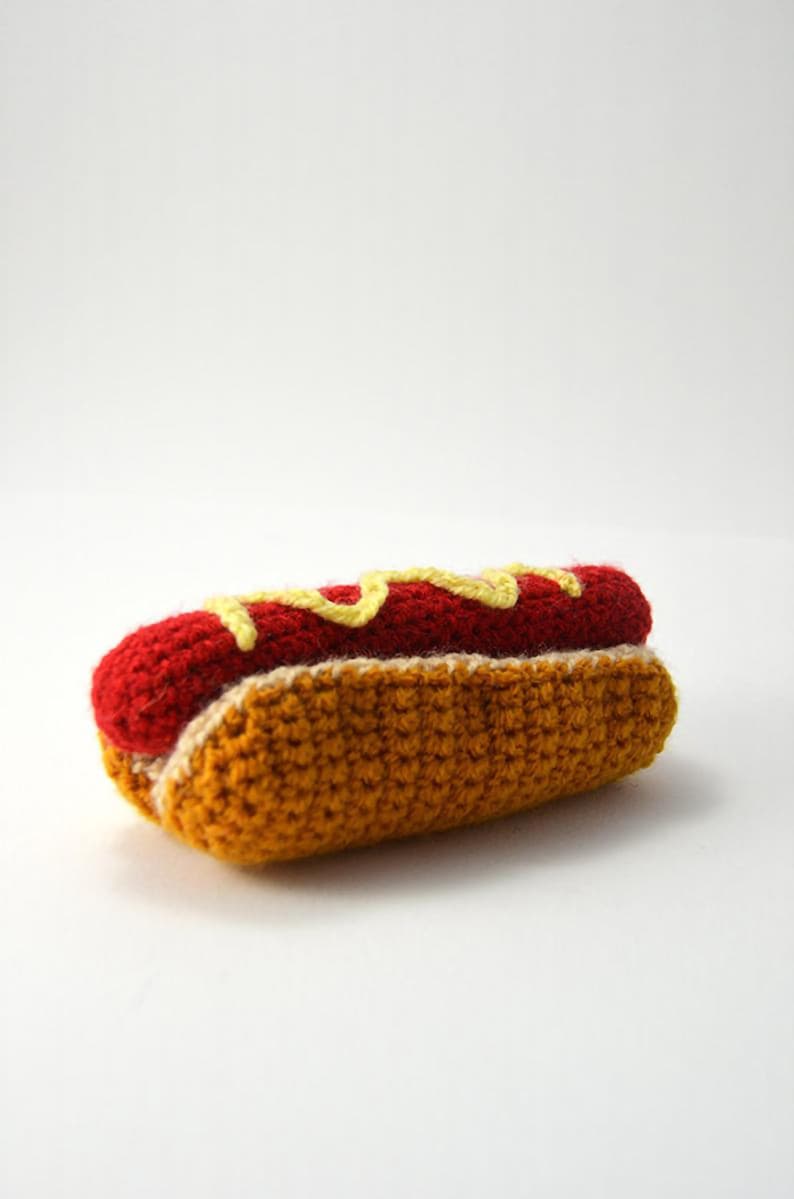 Op de afbeelding: Een gehaakte hotdog met gele mosterd en ketchup. De hotdog is gemaakt van rood garen en het broodje is gemaakt van bruin garen.