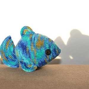 May include: A crocheted blue, green, and gold fish toy with a black eye. The fish is sitting on a brown surface.