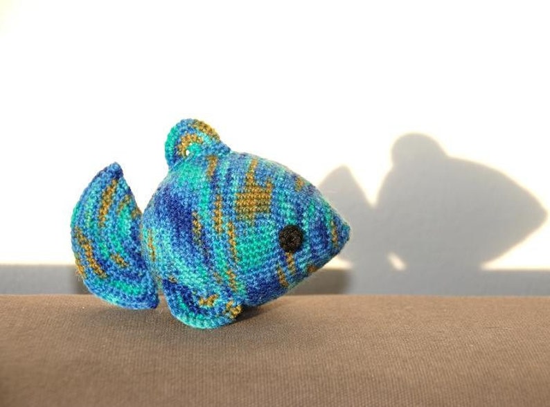 May include: A crocheted blue, green, and gold fish toy with a black eye. The fish is sitting on a brown surface.