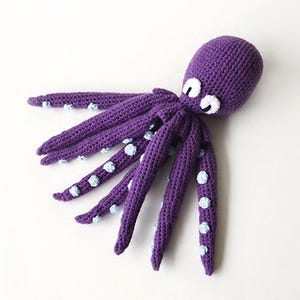May include: A handmade, purple crochet octopus with white eyes and light blue accents on its tentacles. The octopus is lying on a white surface. This soft toy is perfect for children.