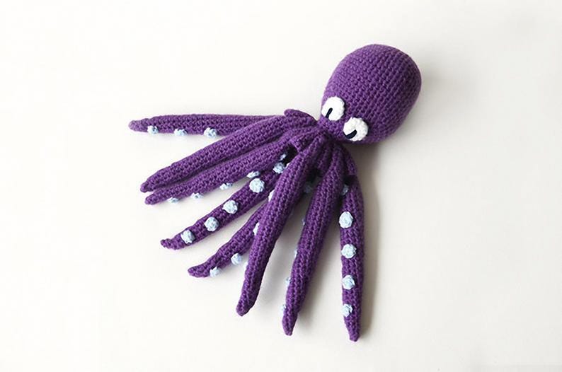 May include: A handmade, purple crochet octopus with white eyes and light blue accents on its tentacles. The octopus is lying on a white surface. This soft toy is perfect for children.