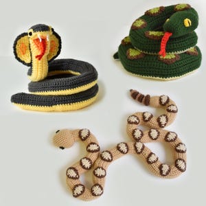 May include: Three crocheted snake toys in different colours. One is a cobra with a yellow hood and a black and yellow body. Another is a green snake with brown and yellow spots. The third is a tan snake with brown and white spots.