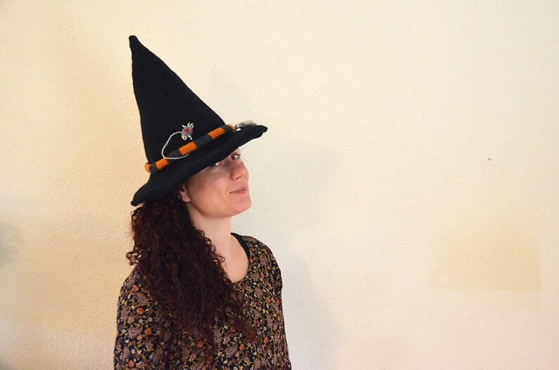 May include: A woman wearing a black witch hat with orange and black trim and a small decorative spider. The hat has a pointed crown and a wide brim.