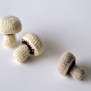 Op de afbeelding: Drie gehaakte paddenstoelen in wit en beige. De paddenstoelen zijn gemaakt van een getextureerd garen en hebben bruine vilt accenten.