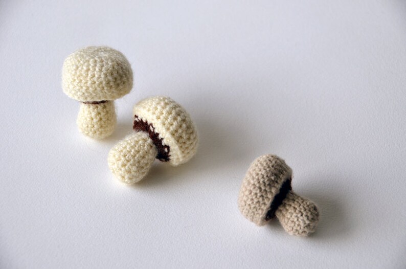 Op de afbeelding: Drie gehaakte paddenstoelen in wit en beige. De paddenstoelen zijn gemaakt van een getextureerd garen en hebben bruine vilt accenten.