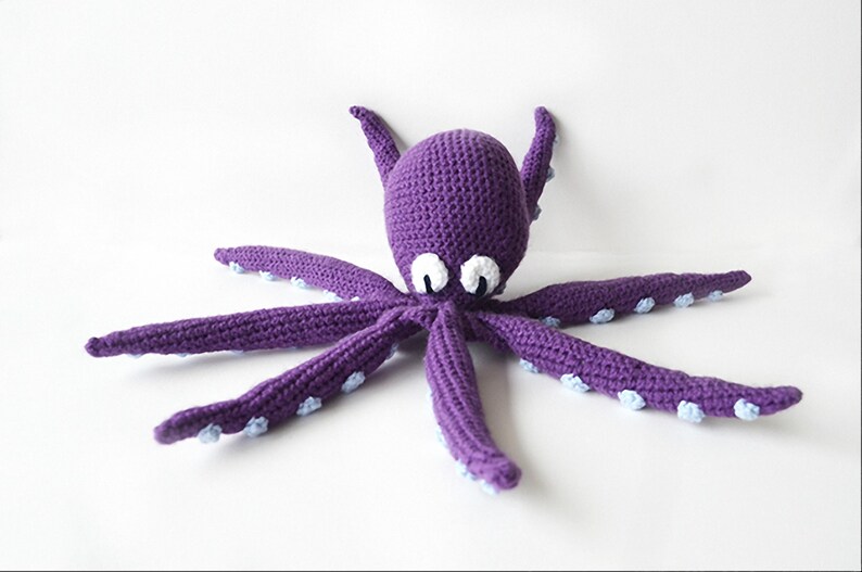 May include: A handmade purple crochet octopus with white and blue accents. The octopus has eight long tentacles and large white eyes. The toy is made of soft yarn and is suitable for children.