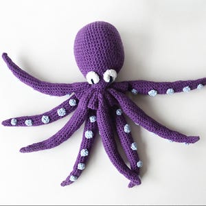 May include: A handmade purple crochet octopus with eight long tentacles. Each tentacle has small, light blue accents. The octopus has white eyes and is laying on a white surface. This plush toy is perfect for children.