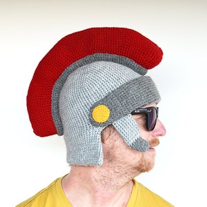 May include: A gray and red crocheted hat shaped like a Roman helmet with a yellow circle on the side. The hat is worn by a person wearing sunglasses.
