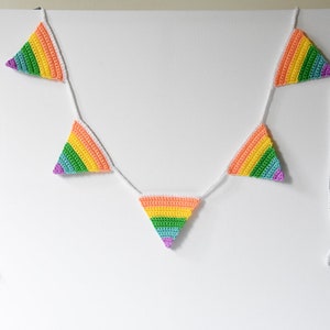 May include: A rainbow crochet banner with triangular flags hanging from white string. The flags are crocheted in a rainbow pattern with purple, blue, green, yellow, orange, and red.