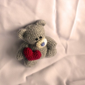 May include: A gray crocheted teddy bear holding a red heart. The bear has a blue button nose and is lying on a white sheet.