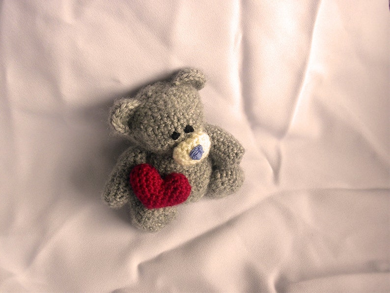 May include: A gray crocheted teddy bear holding a red heart. The bear has a blue button nose and is lying on a white sheet.