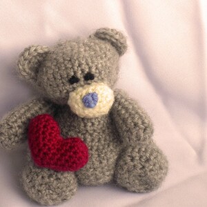May include: A gray crocheted teddy bear holding a red heart. The bear has a blue nose and white fur around the nose.