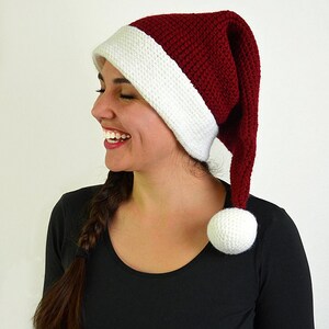May include: A knitted Santa hat with a white brim and a red crown. The hat has a long, pointed end and a white pom-pom. The hat is worn by a person wearing a black long-sleeved shirt.