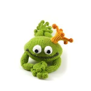 Op de afbeelding: Een groene gehaakte kikker met een oranje gehaakte kroon. De kikker heeft witte ogen en een kleine glimlach.