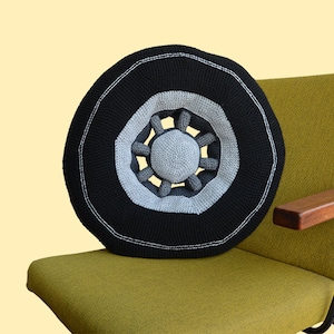 May include: A black and gray crocheted tire pillow with a white center. The pillow is sitting on a green chair with a wooden armrest.