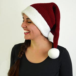 May include: A knitted Santa hat with a white brim and a white pom-pom. The hat is a deep red color. The hat is worn by a person wearing a black long-sleeved shirt.