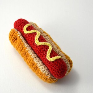 Op de afbeelding: Een gehaakte hotdog met gele mosterd. De hotdog is rood en het broodje is bruin en beige.