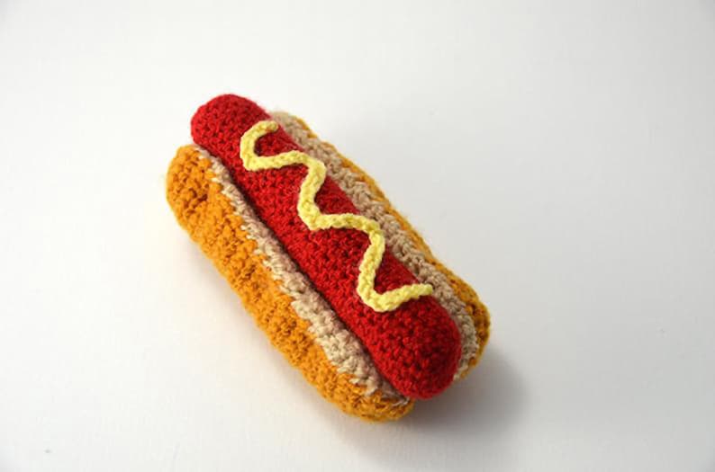 Op de afbeelding: Een gehaakte hotdog met gele mosterd. De hotdog is rood en het broodje is bruin en beige.