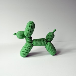 May include: A handmade, green crocheted dog-shaped sculpture. The dog is made of individual, rounded sections, resembling a balloon animal. The sculpture is displayed on a white surface against a plain background.