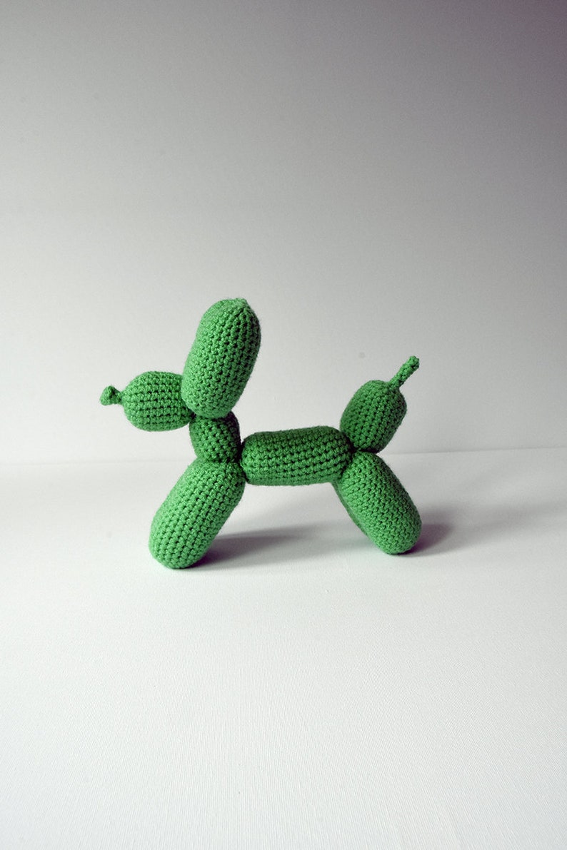 May include: A handmade, green crocheted dog-shaped sculpture. The dog is made of individual, rounded sections, resembling a balloon animal. The sculpture is displayed on a white surface against a plain background.