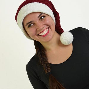 May include: A knitted Santa hat with a burgundy crown and a white band and pom-pom. The hat is worn by a person wearing a black long-sleeved shirt. The person is smiling.