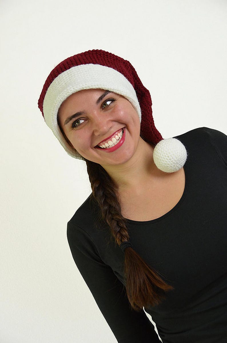 May include: A knitted Santa hat with a burgundy crown and a white band and pom-pom. The hat is worn by a person wearing a black long-sleeved shirt. The person is smiling.