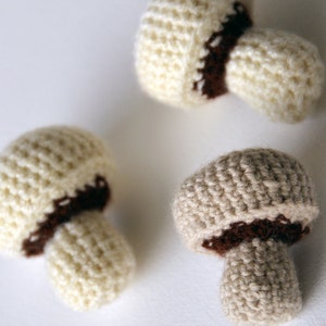 Op de afbeelding: Drie gehaakte paddenstoelen in verschillende tinten beige en bruin. De paddenstoelen hebben een textuur en een bruine band rond het midden.