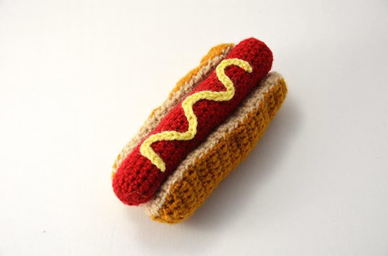 Op de afbeelding: Een gehaakte hotdog met mosterd, gemaakt van rood, geel en bruin garen.