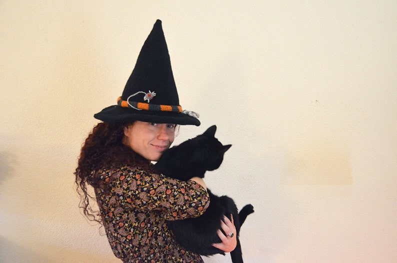 May include: A woman wearing a black witch hat with orange and grey trim holds a black cat in her arms.