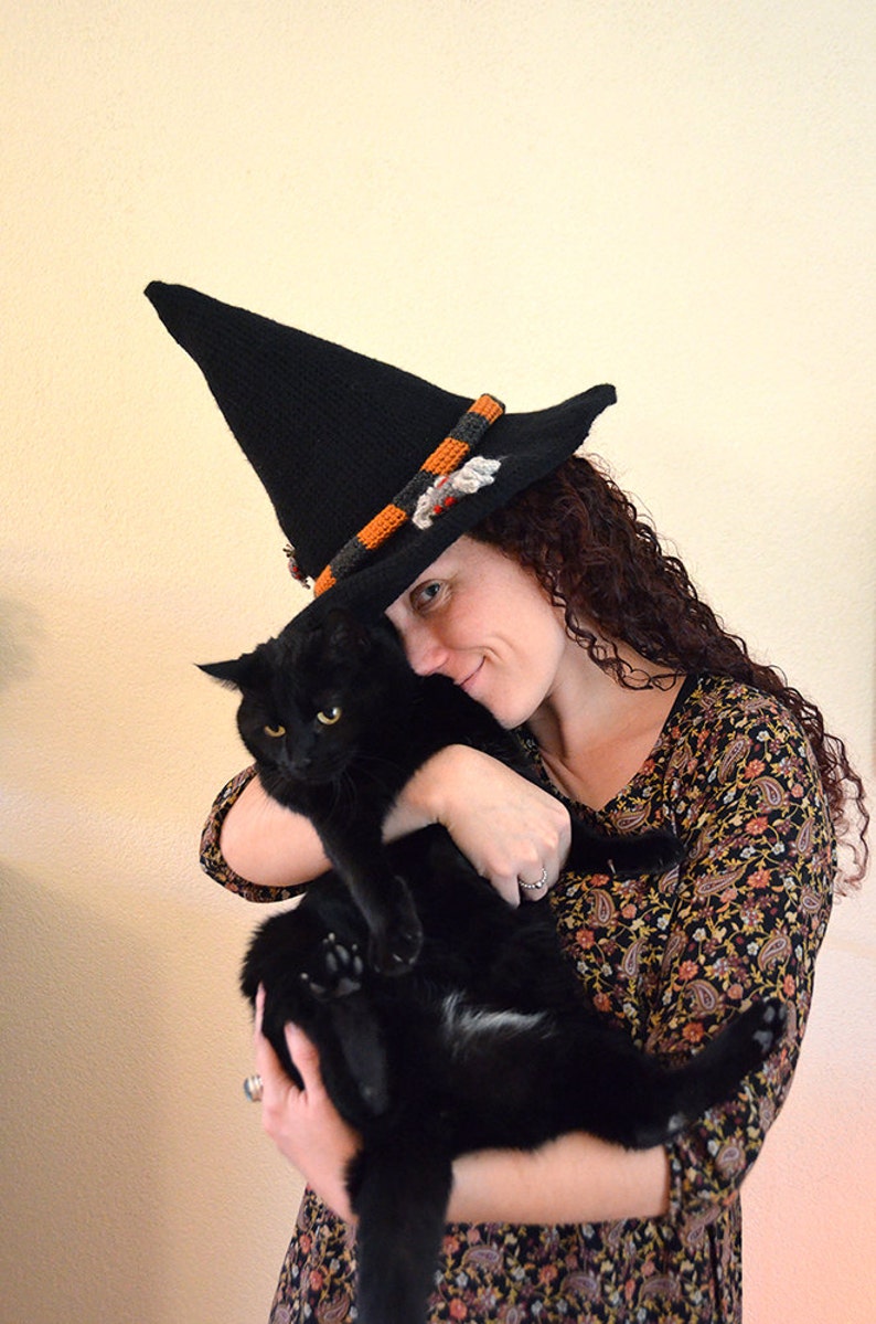 May include: A woman wearing a black witch hat with orange and gray trim holds a black cat in her arms. She is smiling and looking at the camera.