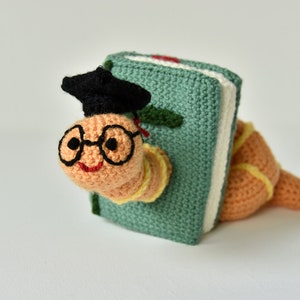 May include: A crocheted worm wearing glasses and a graduation cap, peeking out from behind a green book.